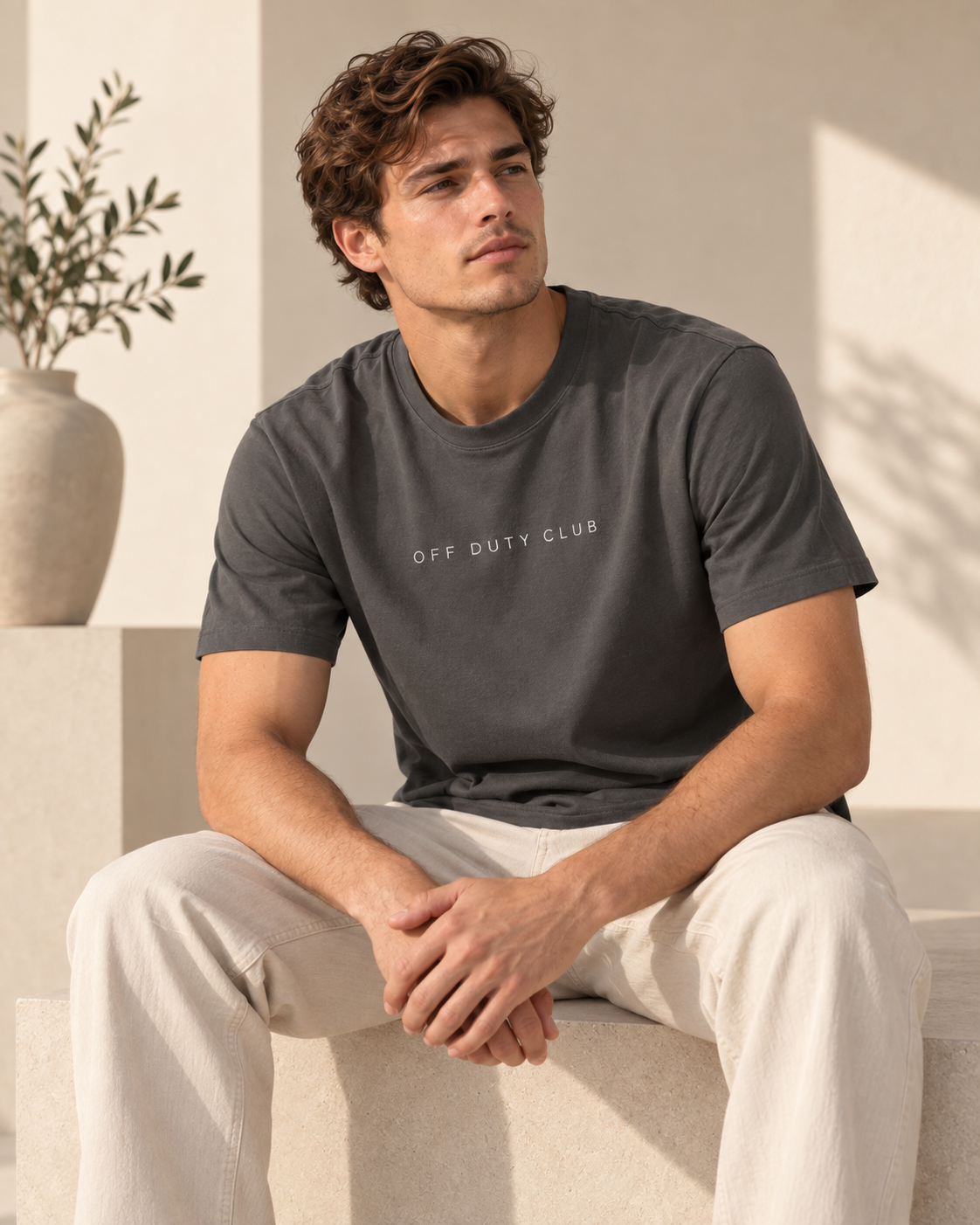 Junger Mann im dunkelgrauen Premium T-Shirt mit minimalistischer „Off Duty Club“-Typografie in warmem, modernem Studio-Look.